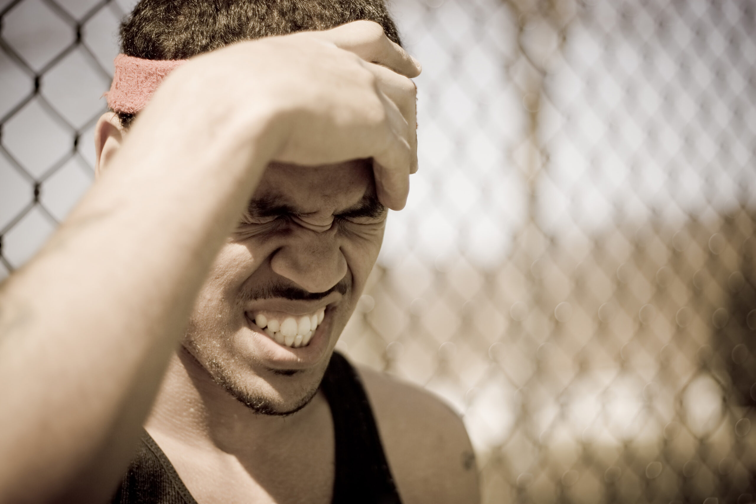 Close-up of a man laughing, hand covering part of his face, wearing a pink sweatband, with a chain-link fence in the background.