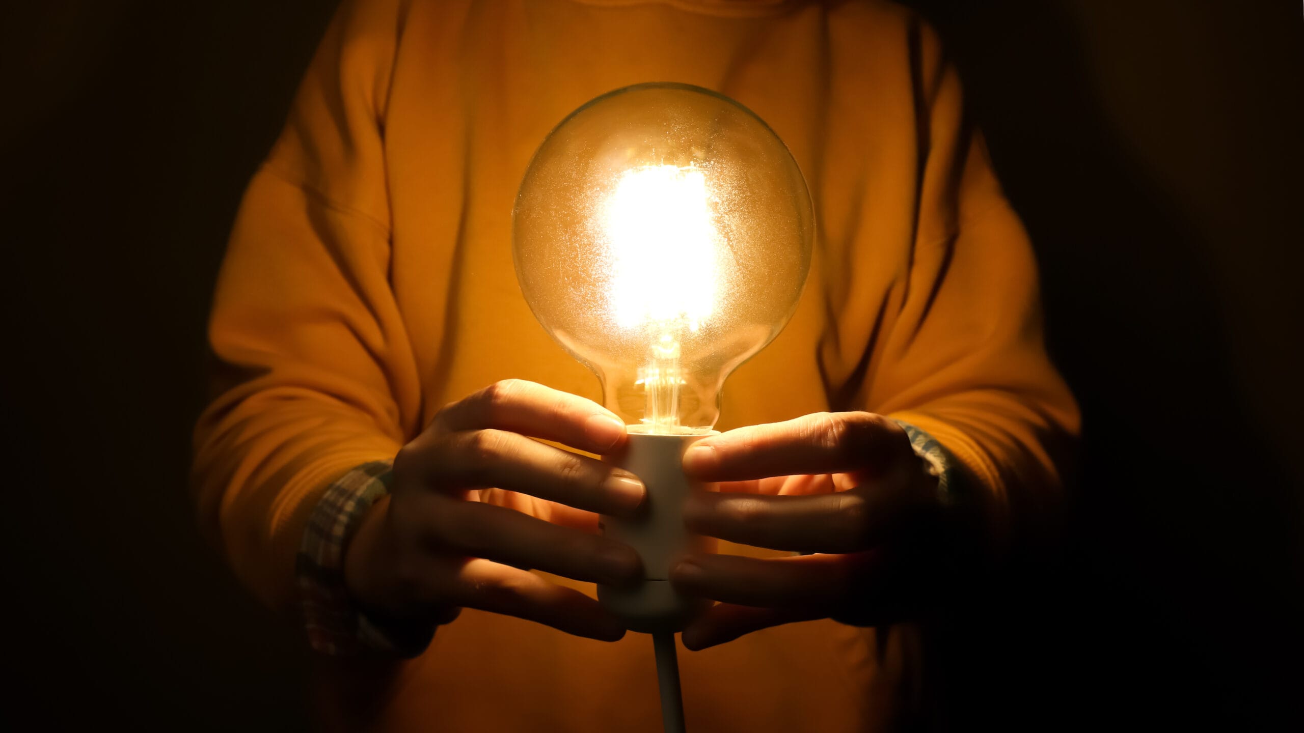 Hands holding glowing light bulb in front of torso on dark background. A pair of hands are gently holding a brightly glowing light bulb, symbolizing a breakthrough solution, a flash of inspiration.