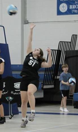 Female volleyball player wearing a black jersey with number 51 jumps to spike the ball in an indoor gym, while a boy with a ball watches in the background.