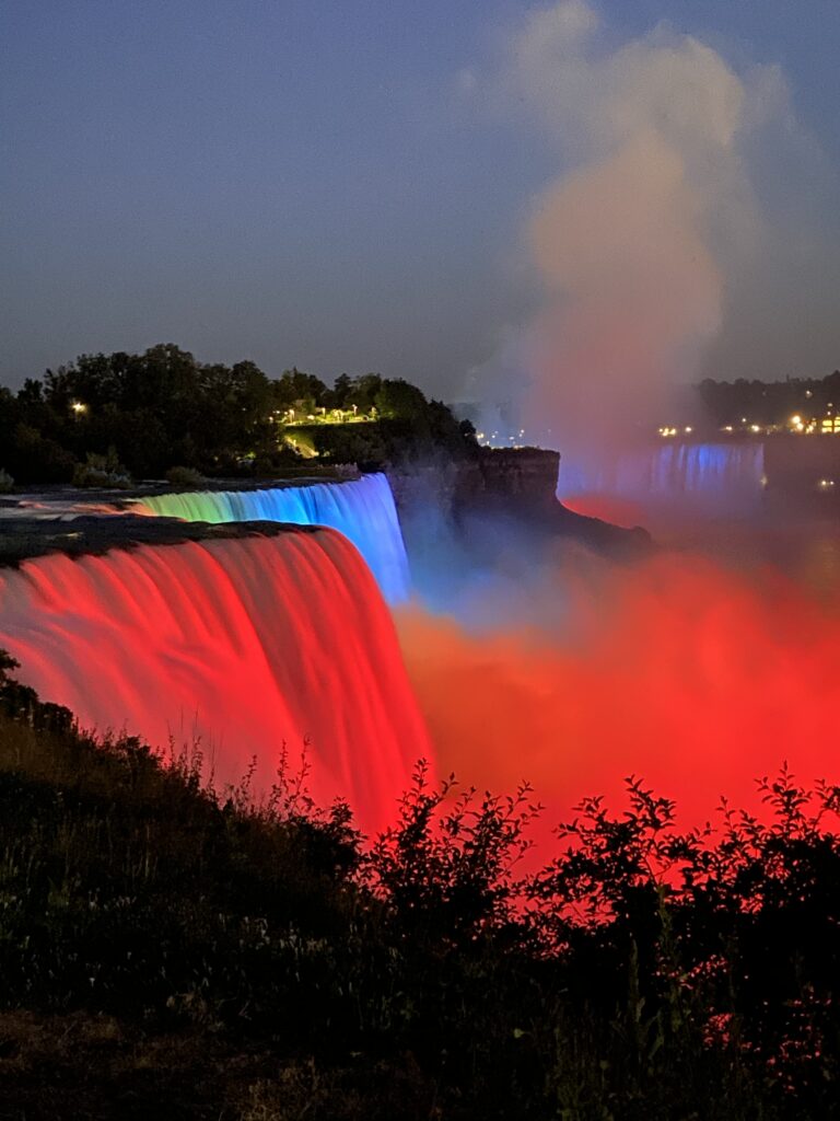 Niagara Falls at night illuminated by rainbow-colored lights with mist rising above the water and trees in the foreground.