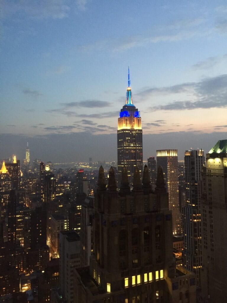 NYC skyline at dusk featuring the Empire State Building lit in blue and yellow against a dusky sky.