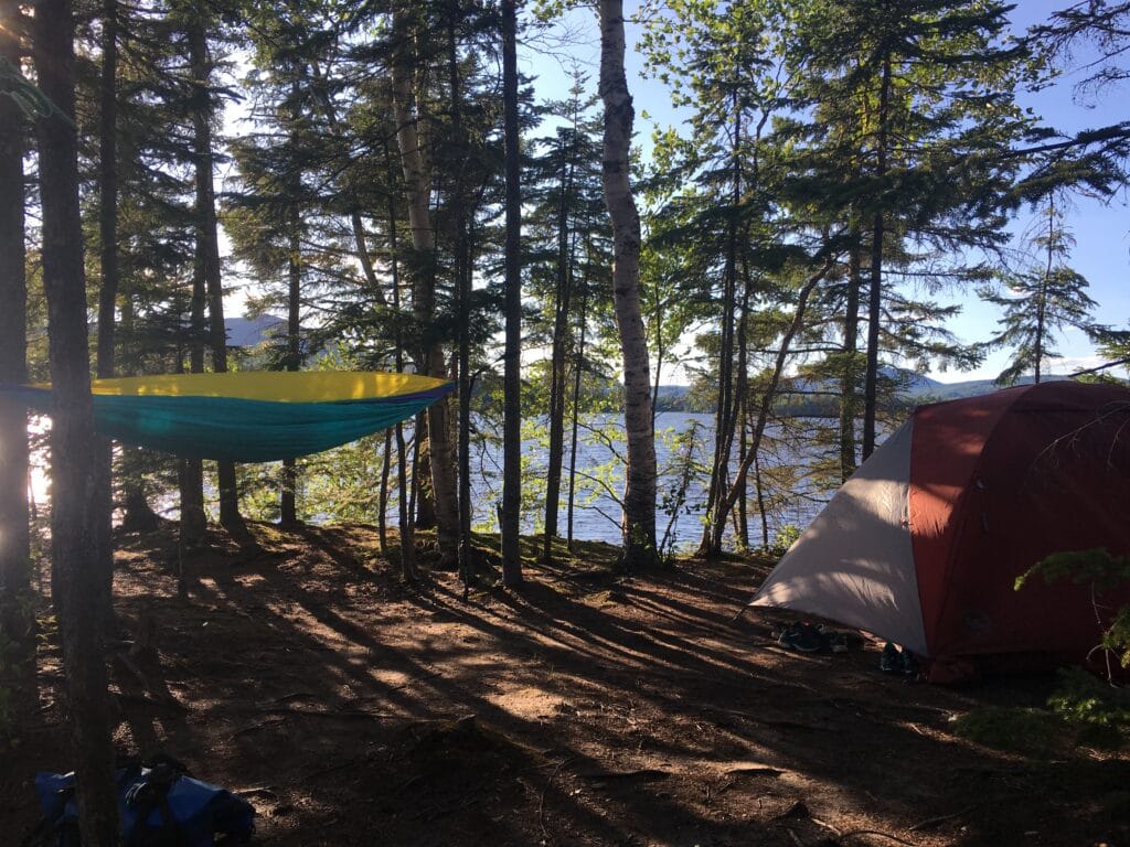 Campsite among tall trees by a lake with a colorful hammock (yellow and teal) and tents nearby, shoes on the ground.