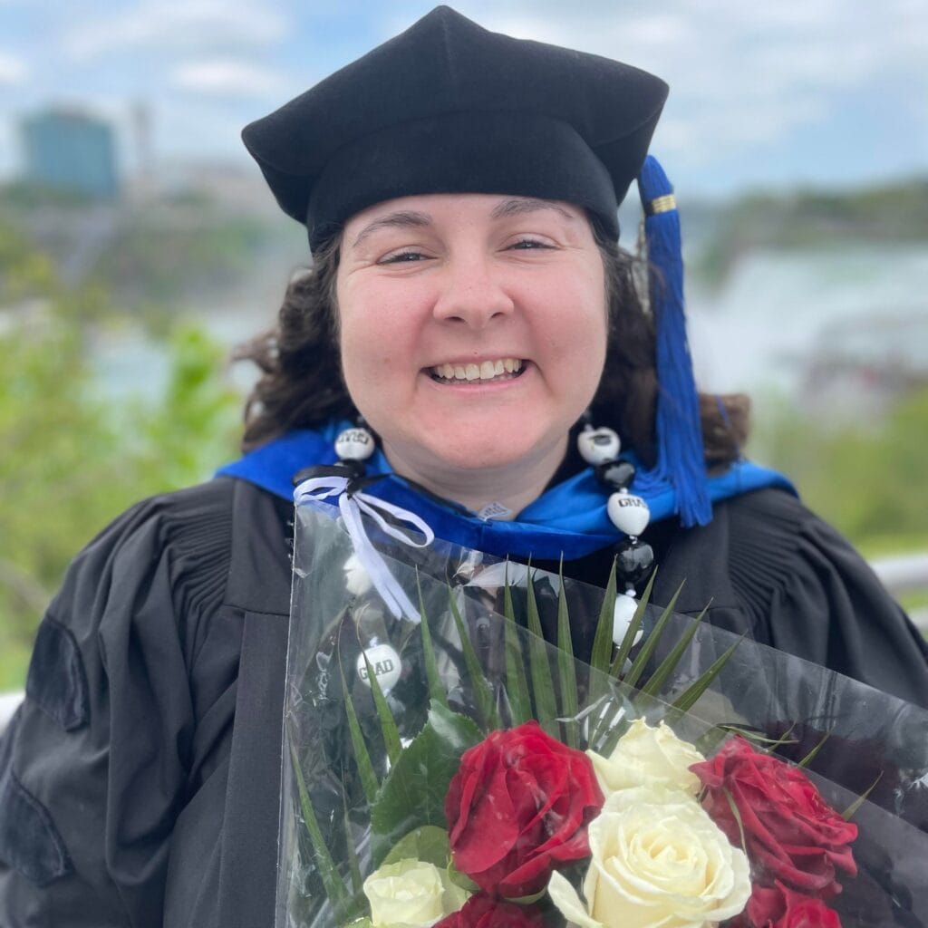 Smiling graduate in a black cap and gown holding a plastic-wrapped bouquet of red and white roses outdoors.