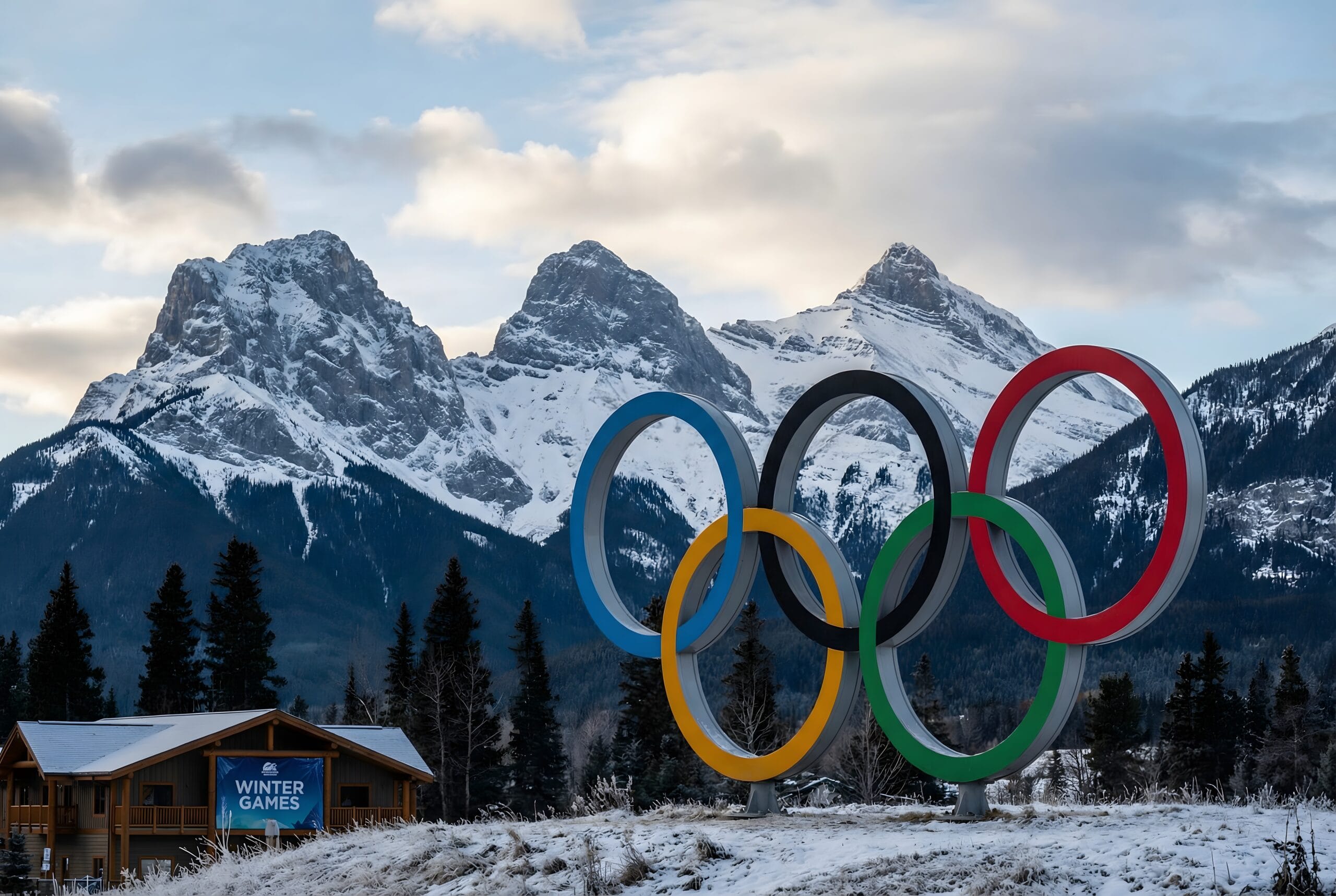 Colorful Olympic rings sculpture in a snowy alpine landscape with a small wooden building displaying a Winter Games banner.