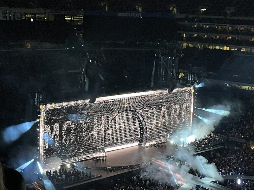 Large concert stage at night with intricate metal rigging and a standing and seated audience in a dark arena.
