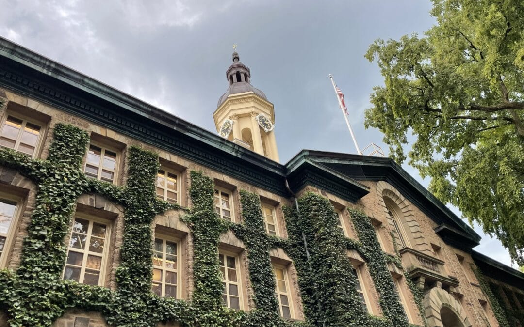 Beige clock tower rising above an ivy-covered stone building, with a flagpole and American flag nearby under a cloudy blue sky.