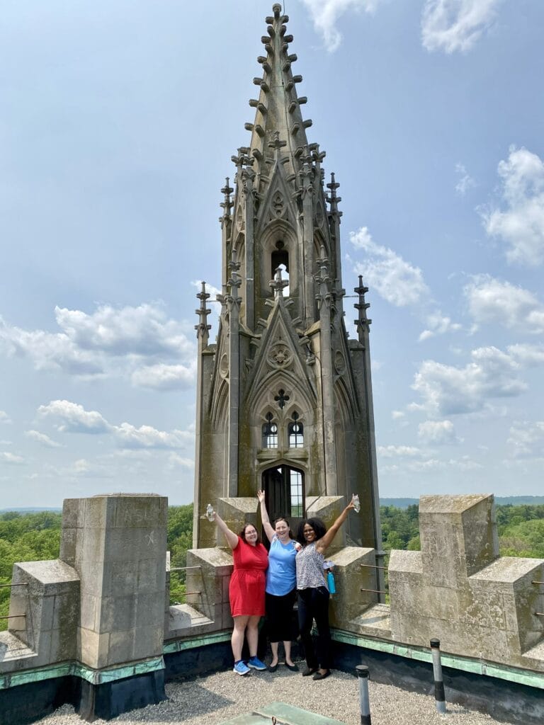 Three women stand on a stone viewing platform at the base of a tall Gothic spire, raising their arms in celebration against a blue sky.