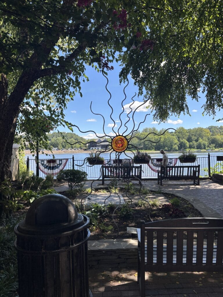 Metal sun sculpture with swirling tentacles near a riverside park bench seating area under trees by a fence and patriotic bunting behind the water.