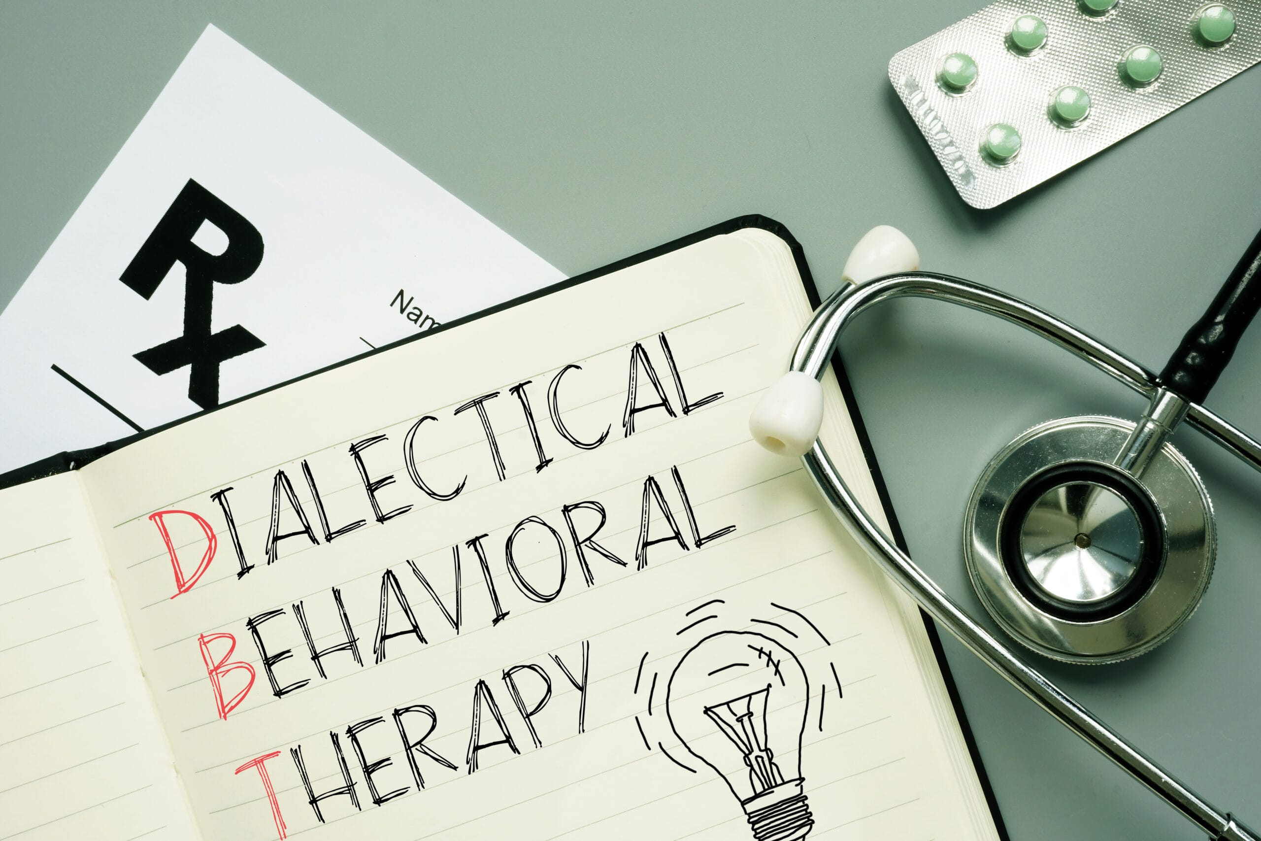 DBT Graphic | Grand Slam Psych Open notebook page with handwritten words including 'DIABETICAL' and 'BEHAVIORAL THERAPY,' beside a stethoscope and a blister-pack of pills on a desk.