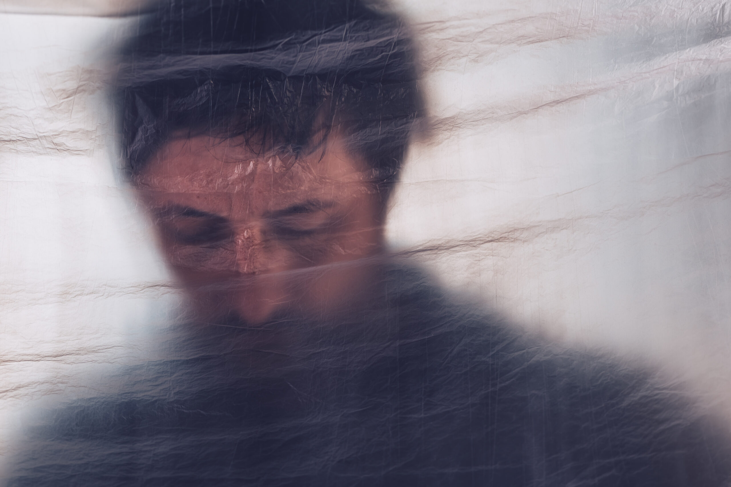 Blurred close-up of a person's eyes and forehead behind a translucent, wrinkled plastic sheet laced with scratches.